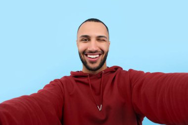 Gülümseyen genç adam açık mavi arka planda selfie çekiyor.