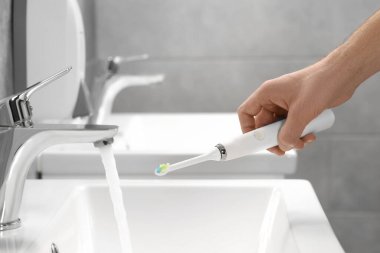 Banyoda lavabonun üzerinde akan suyun yanında elektrikli diş fırçası tutan adam, yakın plan.