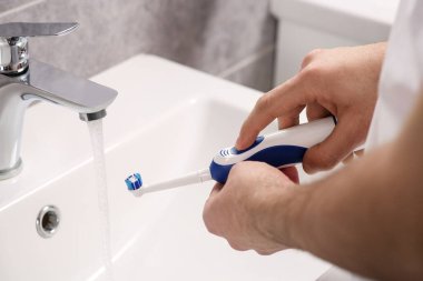 Banyoda lavabonun üzerinde akan suyun yanında elektrikli diş fırçası tutan adam, yakın plan.