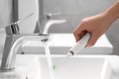Banyoda lavabonun üzerinde akan suyun yanında elektrikli diş fırçası tutan adam, yakın plan.