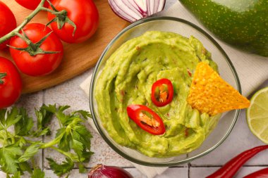 Bir kase lezzetli guacamole, cips ve malzemeler beyaz fayanslı masada, düz yerde.