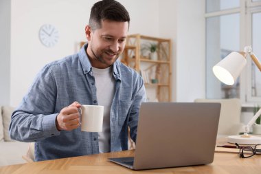 İçkili mutlu bir adam odasındaki ahşap masada dizüstü bilgisayarla çalışıyor.