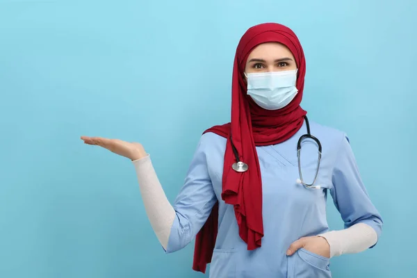 头戴头巾 身穿医疗制服 戴着防护面具的穆斯林妇女指着浅蓝色背景的东西 案文的篇幅 — 图库照片