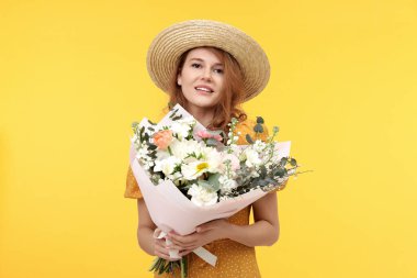 Sarı arka planda bir buket güzel çiçekle saman şapkalı mutlu kadın.