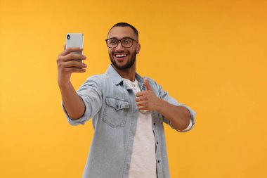 Gülümseyen genç adam akıllı telefonuyla selfie çekiyor ve sarı arka planda baş parmak gösteriyor.