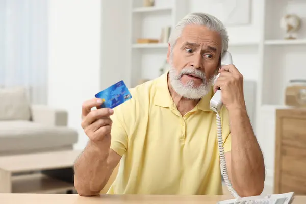 Confused senior man with credit card talking on phone at home. Be careful - fraud