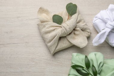 Furoshiki tekniği. Hediyeler farklı kumaşlarda paketlenmiş ve ahşap masada bitkilerle süslenmiş, düz yerde metin için yer var.