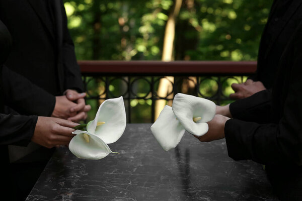 People with calla lily flowers near granite tombstone at cemetery outdoors, closeup. Funeral ceremony