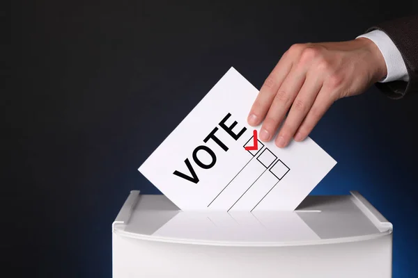 ダークブルーの背景に紙を投票して投票箱に入れる男 — ストック写真