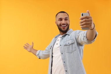 Gülümseyen genç adam sarı arka planda akıllı telefonuyla selfie çekiyor.