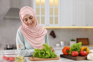 Müslüman kadın, mutfaktaki beyaz masada sebzelerle lezzetli salata yapıyor. Metin için boşluk