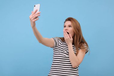 Şok olmuş bir kadın açık mavi arka planda selfie çekiyor.
