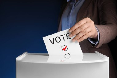 Woman putting paper with word Vote and tick into ballot box on dark blue background clipart