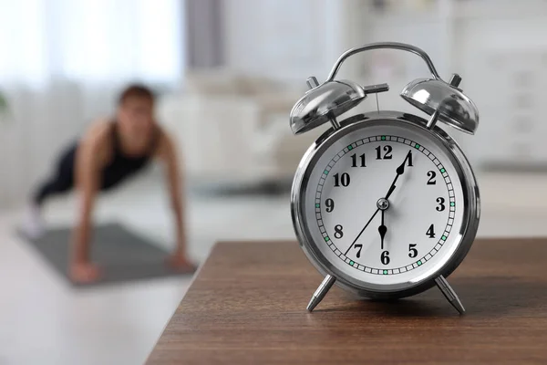 Morning routine. Alarm clock on wooden table and woman doing exercise, selective focus. Space for text