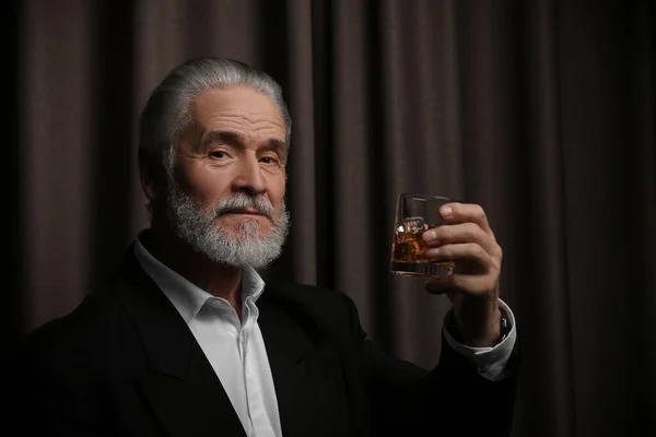 Senior man in formal suit holding glass of whiskey with ice cubes on brown background