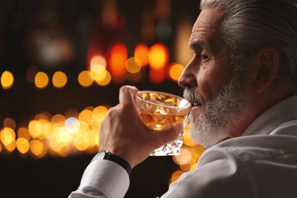 Senior man drinking whiskey against blurred lights, closeup