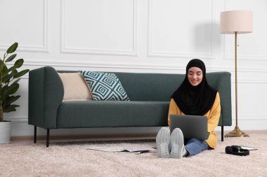 Tesettürlü Müslüman bir kadın odada koltuğun yanında dizüstü bilgisayar kullanıyor. Metin için boşluk
