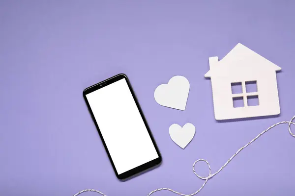 Long-distance relationship concept. Smartphone, white house, paper hearts and decorative cord on violet background, flat lay