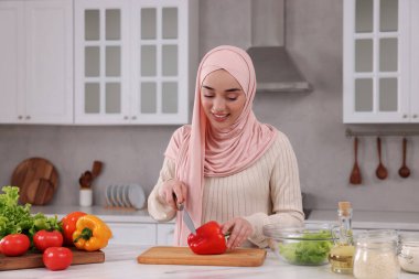 Müslüman kadın, mutfaktaki beyaz masada sebzelerle lezzetli salata yapıyor.