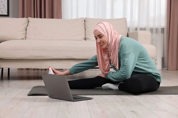Muslim woman in hijab stretching near laptop on fitness mat at home