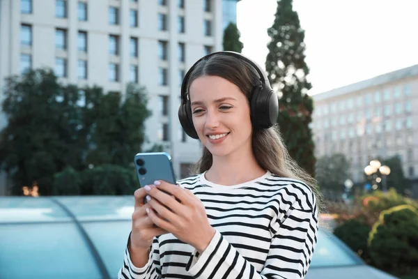 在城市街道上用智能手机对着耳机微笑的女人 — 图库照片