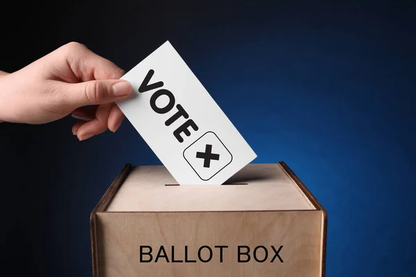 Woman putting paper with word Vote and tick into ballot box on dark blue background