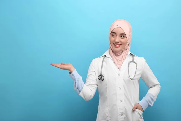 穿着头巾和医疗制服的穆斯林妇女指着浅蓝色背景的东西 — 图库照片