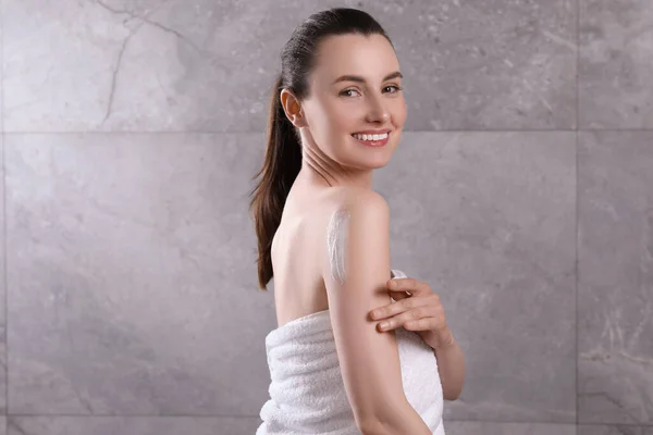 Happy woman applying body cream onto arm near grey wall