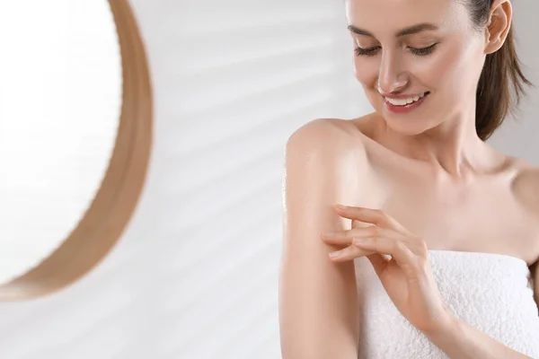 Happy woman applying body oil onto arm in bathroom, space for text