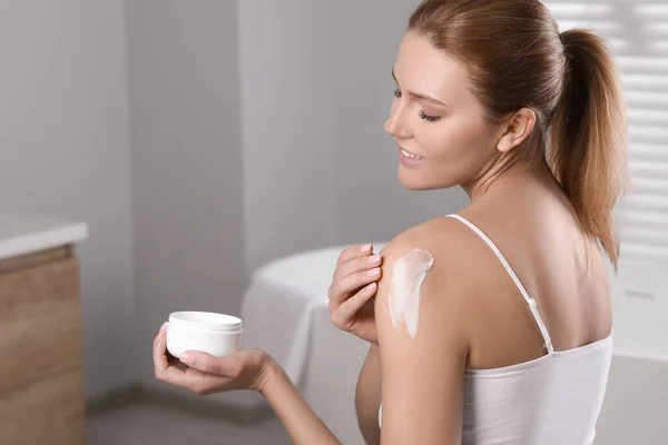 Happy woman applying body cream onto shoulder in bathroom, space for text