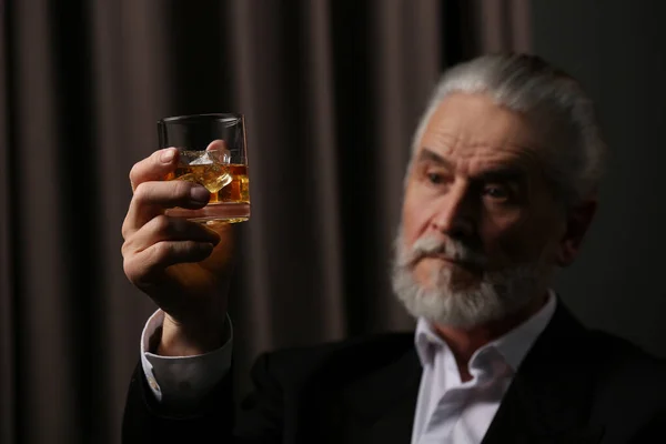 Senior man in suit holding glass of whiskey with ice cubes on brown background, selective focus