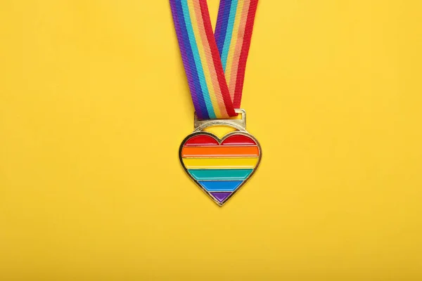 Rainbow ribbon with heart pendant on yellow background, top view. LGBT pride