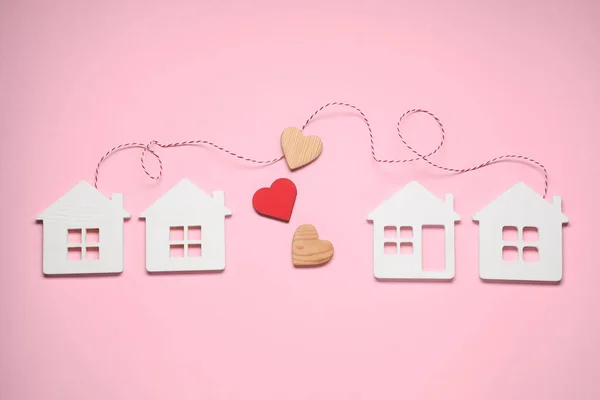 Decorative hearts and cord between white house models on pink background symbolizing connection in long-distance relationship, flat lay