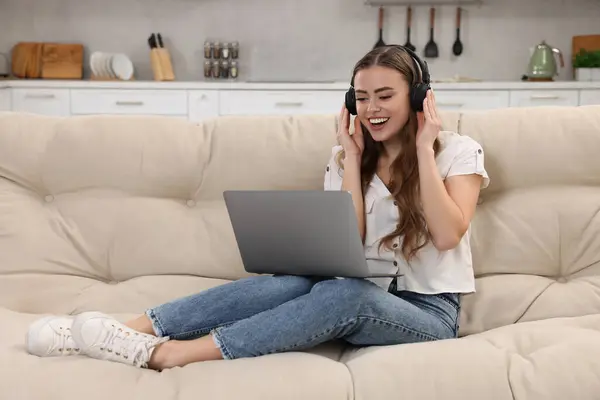 Happy woman with laptop listening to music in headphones on couch