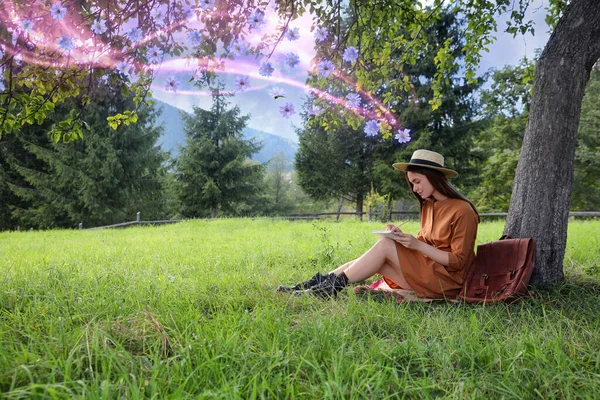 緑の草の上の木の近くのノートパッドの鉛筆で描く美しい若い女性 彼女の象徴的なマインドフルネスの状態の近くを飛ぶ花 — ストック写真