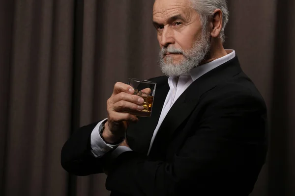 Senior man in suit holding glass of whiskey with ice cubes on brown background. Space for text