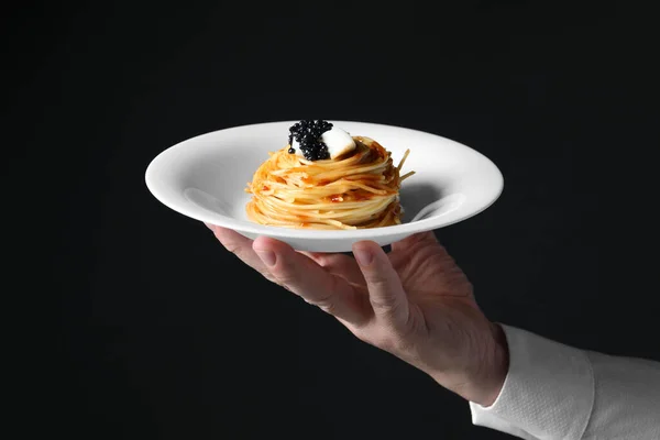 Waiter holding plate of tasty spaghetti with tomato sauce and black caviar on dark background, closeup. Exquisite presentation of pasta dish