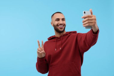 Gülümseyen genç adam akıllı telefonuyla selfie çekiyor ve açık mavi arka planda barış işareti gösteriyor.