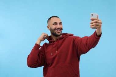 Gülümseyen genç adam açık mavi arka planda akıllı telefonuyla selfie çekiyor.