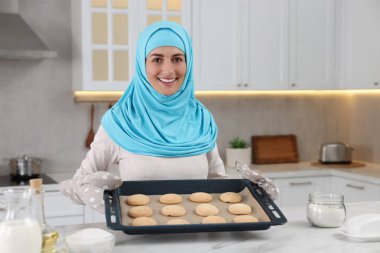 Mutfaktaki beyaz masada elinde kurabiyelerle Müslüman bir kadının portresi.