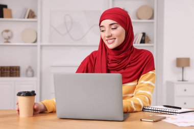 Tesettürlü Müslüman kadın odasındaki tahta masada dizüstü bilgisayar kullanıyor.