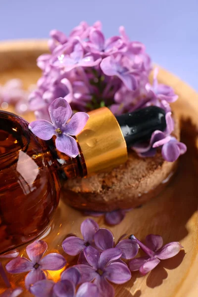 Bottle with essential oil, lilac flowers and water in bowl on violet background, closeup