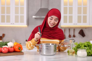 Müslüman kadın, mutfaktaki beyaz masada sebzelerle lezzetli çorba yapıyor.