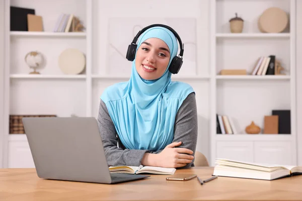 Muslim woman in hijab using laptop at wooden table in room