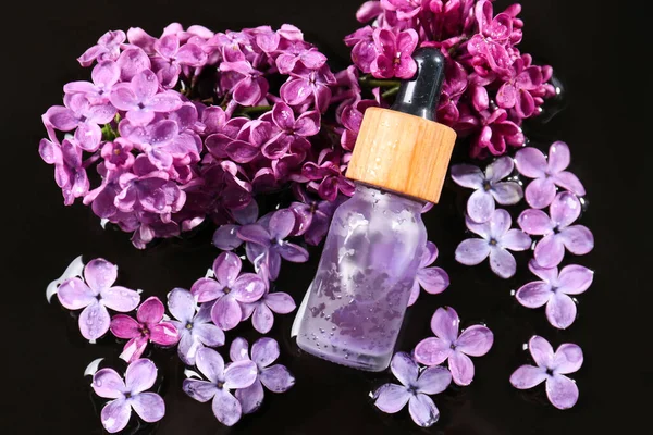 Bottle with essential oil, lilac flowers and water on black surface, flat lay