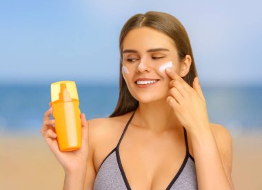 Sun protection. Beautiful young woman applying sunblock onto face on beach clipart