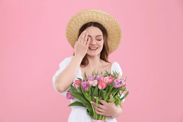 ピンクの背景に美しいチューリップの花束を保持するわら帽子で幸せな若い女性 — ストック写真
