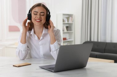 Kulaklıklı mutlu kadın beyaz masada dizüstü bilgisayarın yanında müzik dinliyor.