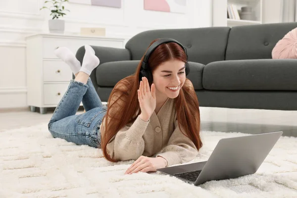 Happy woman with laptop and headphones having video chat on rug in living room