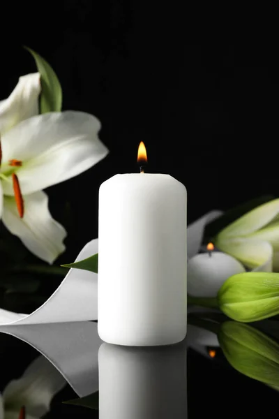 White lilies and burning candle on black mirror surface in darkness, space for text. Funeral symbols